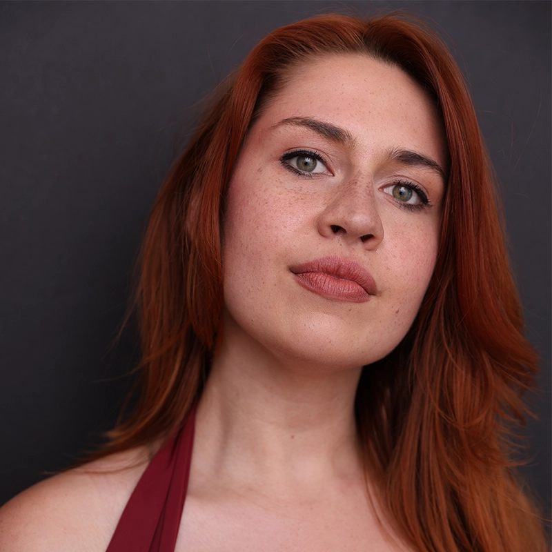 A person with long auburn hair, light skin, and green eyes gazes confidently at the camera. Reminiscent of Kelsey Valente, they wear natural makeup, a maroon top, and are set against a plain dark background. The expression is calm and assertive.
