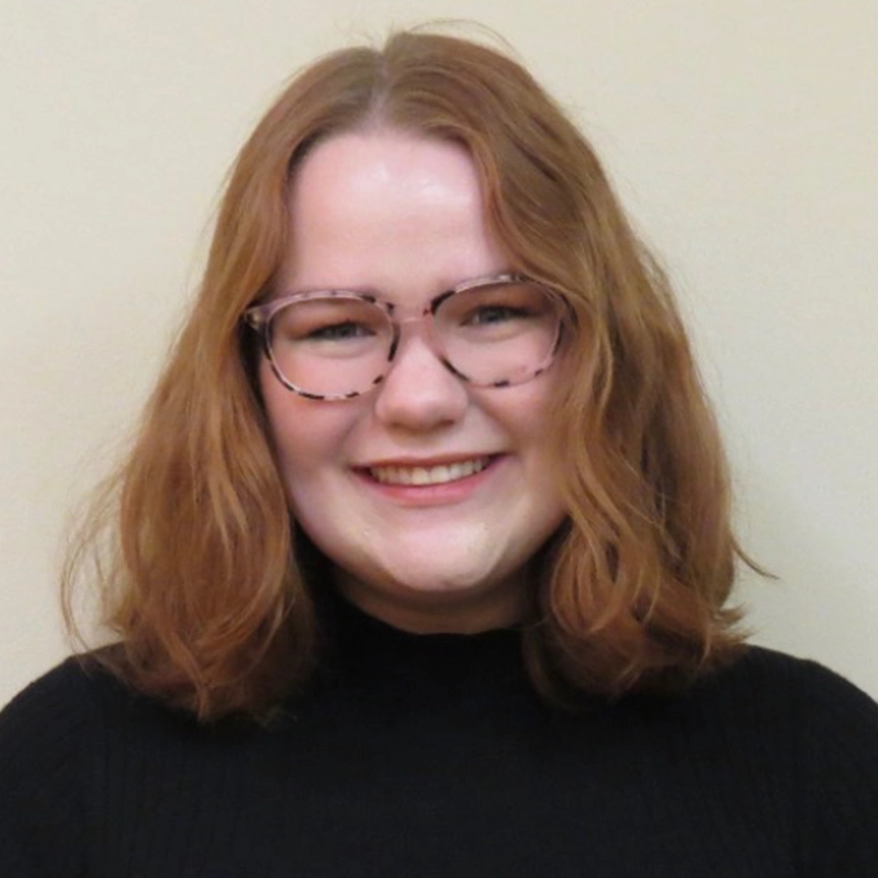 Kendall Tubbs, a young person with shoulder-length wavy red hair and tortoiseshell glasses, smiles at the camera. They wear a black sweater and are in front of a plain, light-colored background.