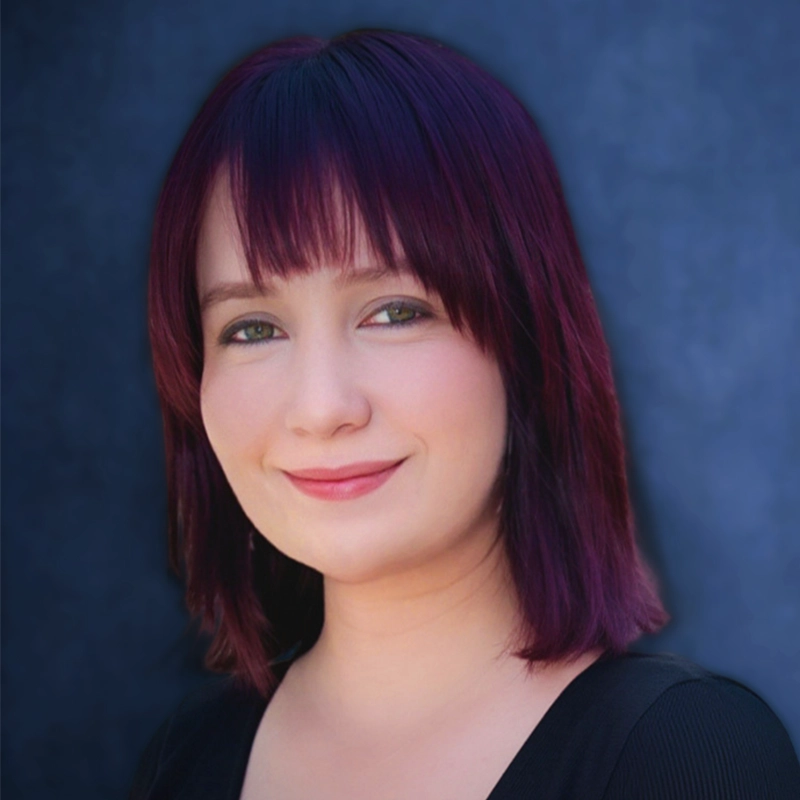 A woman with chin-length, straight, dark purple hair and bangs smiles softly. She wears a black top and has fair skin. The background is a smooth, dark blue gradient. The lighting is even, highlighting her face gently—an image reminiscent of Kendall Tubbs’ understated elegance.