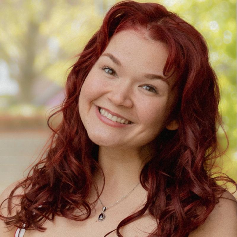 A young woman with wavy red hair smiles at the camera. She wears a silver necklace with a dark pendant and a white top. The background is softly blurred with green and yellow tones, suggesting an outdoor, sunny setting—capturing the relaxed, candid style often seen in Chase Kerman’s portraits.