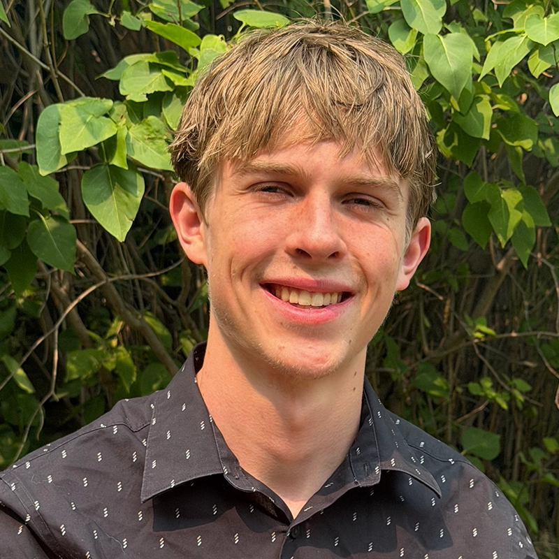 A young person with light brown hair and fair skin smiles outdoors. They wear a black button-up shirt with a subtle white pattern. Green leaves and branches fill the background, indicating a natural, sunny setting.