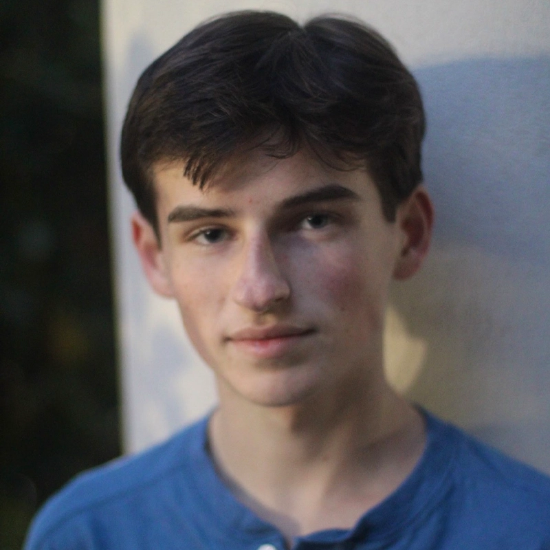 A teenage boy with short brown hair and fair skin stands in soft focus against a light wall outdoors. He wears a blue henley shirt and has a neutral expression. The background is blurred, highlighting his face and upper torso—this portrait captures Kevin Kirley in a quiet, contemplative moment.