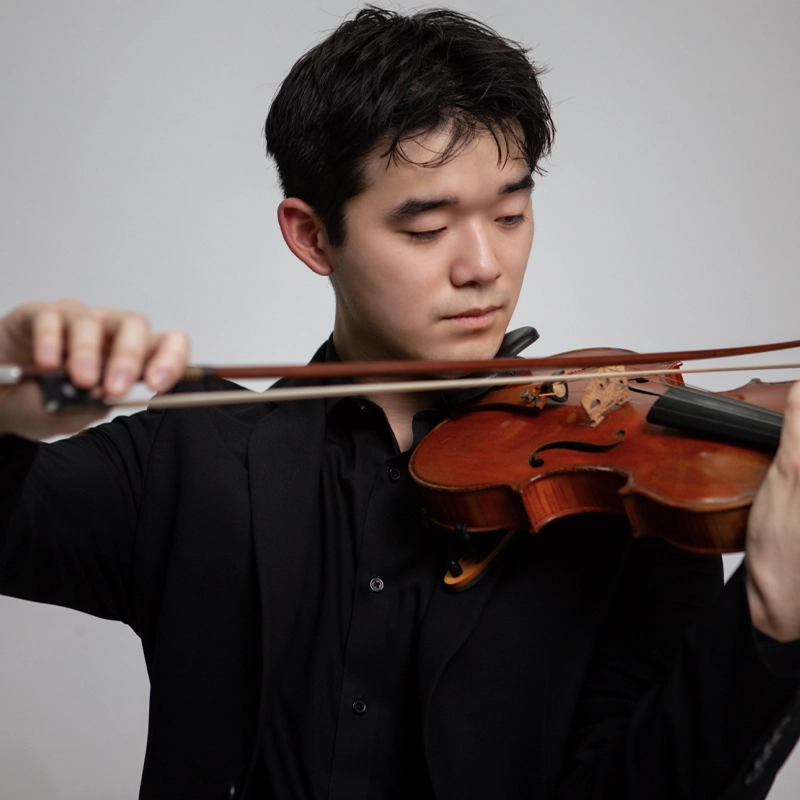 A young man in a black suit, identified as Riku Korenaga, plays a violin, focusing intently. He holds the bow in his right hand and supports the violin with his left hand. The background is plain and light-colored, drawing attention to his expression and the instrument.