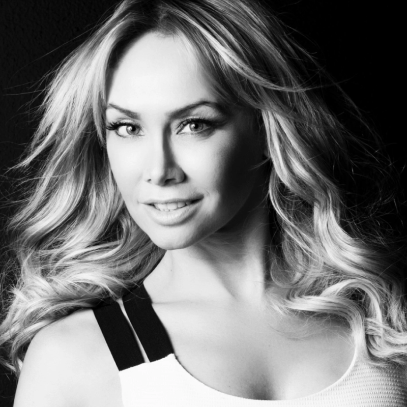 Black and white portrait of a woman with long, wavy blond hair, light eyes, and subtle makeup. She is smiling slightly, facing the camera, and wearing a sleeveless top with dark straps against a dark background.