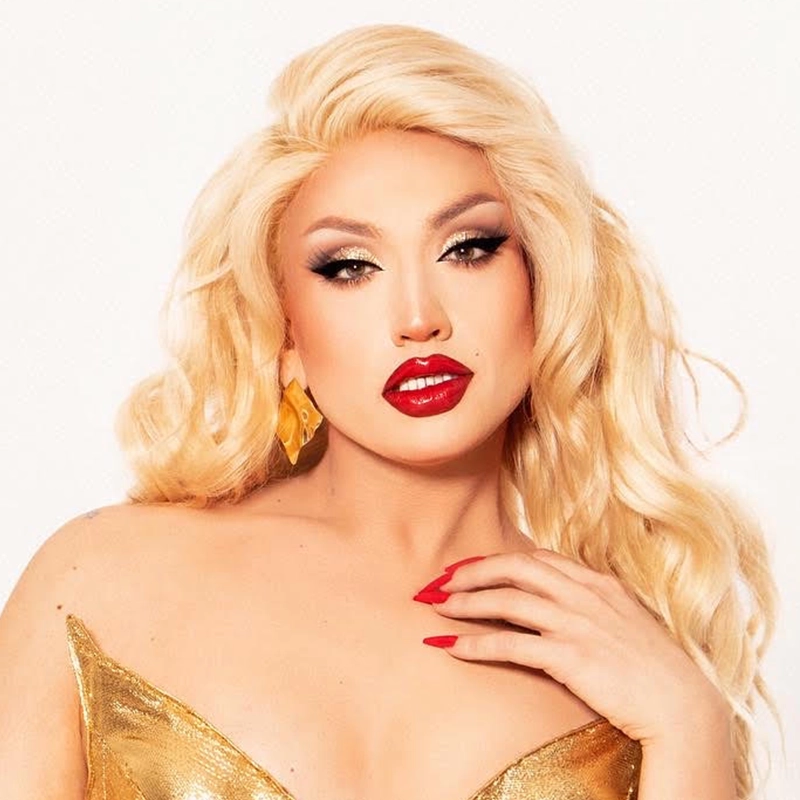 A person with long, wavy platinum blonde hair poses in a glamorous gold dress. Channeling John Arthur Hill’s signature flair, they have dramatic winged eyeliner, bold red lipstick, and red nails. Large gold earrings and flawless makeup create a striking, confident look against a plain white background.