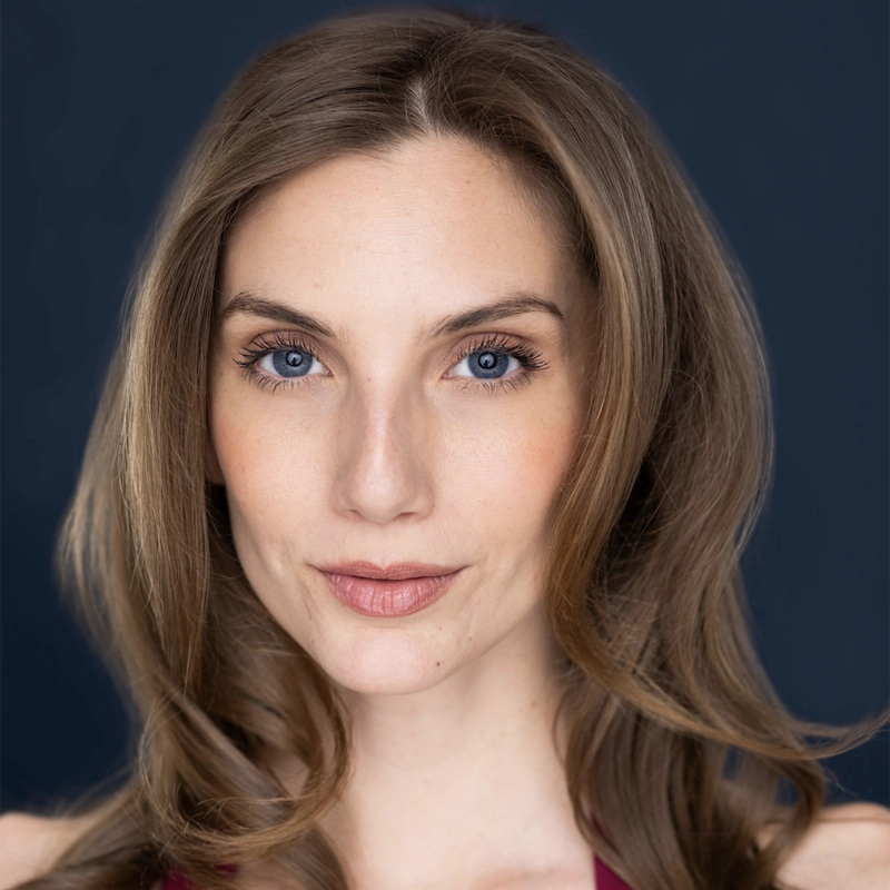 A woman with long light brown hair and blue eyes looks directly at the camera, smiling softly. She has fair skin, natural makeup, and wears a sleeveless maroon top. The background is a solid dark blue. The lighting is even and bright, highlighting the calm confidence of Amy Laviolette.