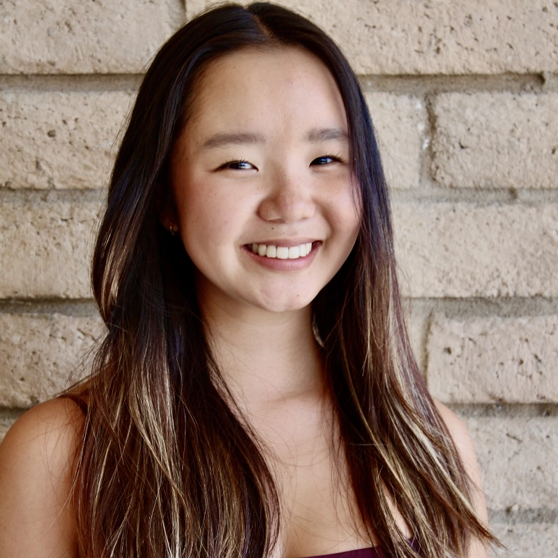 A woman with long, straight dark hair and lighter highlights smiles at the camera. She is wearing a sleeveless top and stands in front of a beige brick wall. The lighting is natural, highlighting her face and hair.