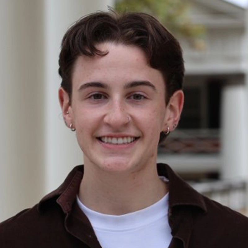 A young person with short brown hair, hoop earrings, and a nose ring smiles outdoors. They wear a brown jacket over a white shirt. The background is blurred, showing light-colored columns and an indistinct building, creating a warm and inviting setting for Celeste Zwingel.