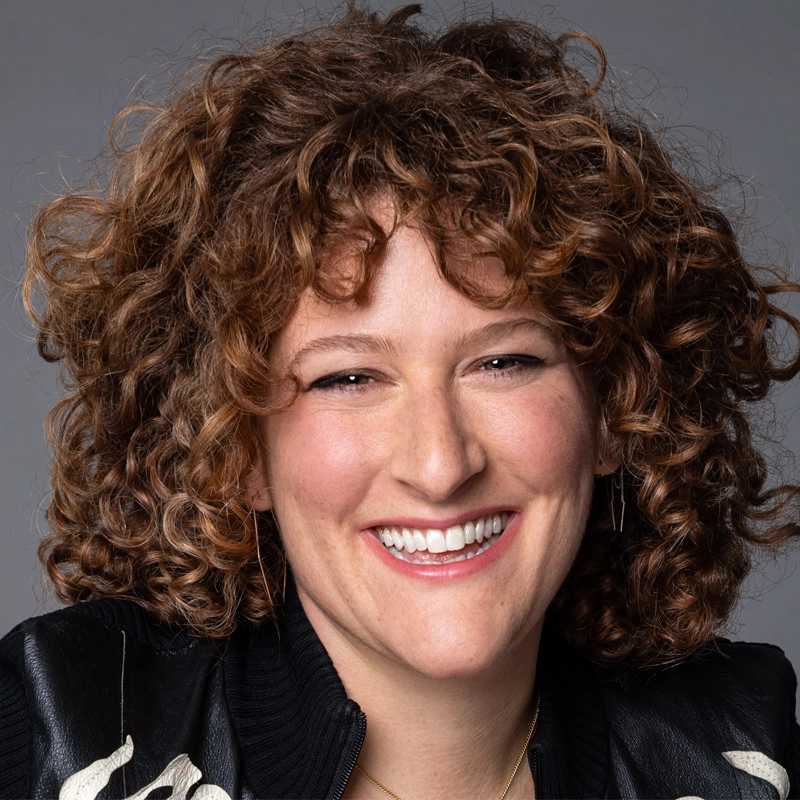 Smiling person with light skin, curly brown hair, wearing a black jacket with white detailing. The background is plain grey. Their expression is friendly and joyful, and they are facing the camera—this cheerful portrait features Samantha Levy.