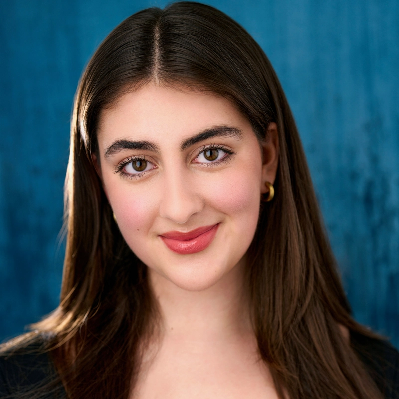 A young woman with long brown hair and light skin smiles gently. She wears gold hoop earrings and a black top. The background is a textured blue, creating a professional, polished headshot look. Her makeup is natural with pink lipstick.
