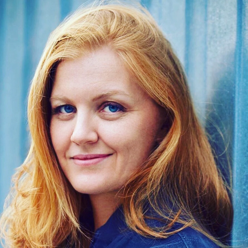 A woman with shoulder-length strawberry-blonde hair and bright blue eyes looks toward the camera. She has a gentle smile and is positioned against a textured blue background.