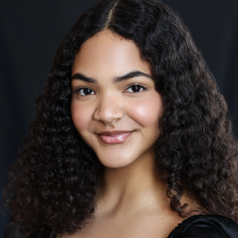Bethany Livers, a young woman with medium brown skin and long, dark, naturally curly hair, smiles gently. She wears a black top and has soft, natural makeup. The background is a plain, dark color, creating a simple, professional portrait.