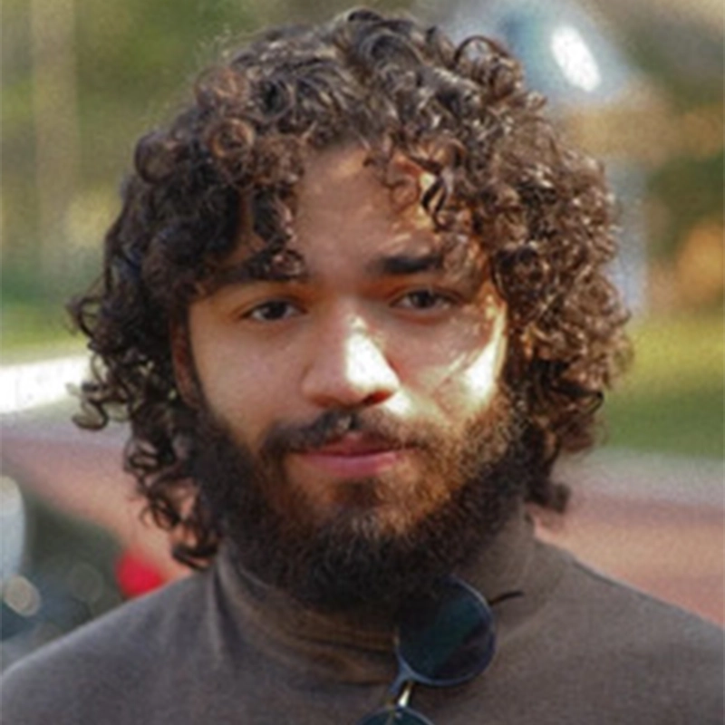 A young person with curly dark hair and a beard looks at the camera, standing outdoors. Salvador Diaz-DeBose wears a brown turtleneck and sunglasses hang from the shirt collar. The background is blurred greenery and walkway, suggesting a park or outdoor setting.