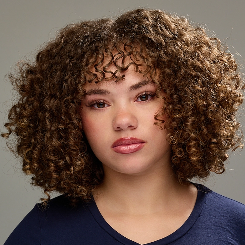 A young person with light brown, tightly curled hair and subtle makeup faces forward. They wear a navy blue shirt and have a calm, neutral expression. The background is plain gray.