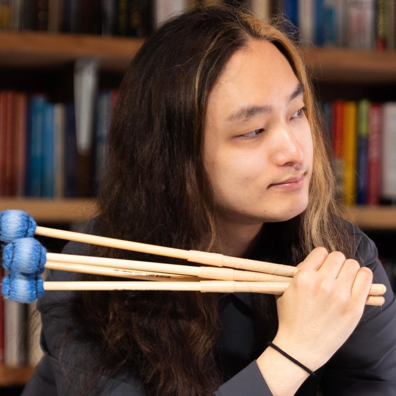 A person with long, dark hair featuring a blond streak holds four blue-tipped mallets over their shoulder and looks to the side. They wear a dark shirt and sit in front of a bookshelf filled with colorful books.