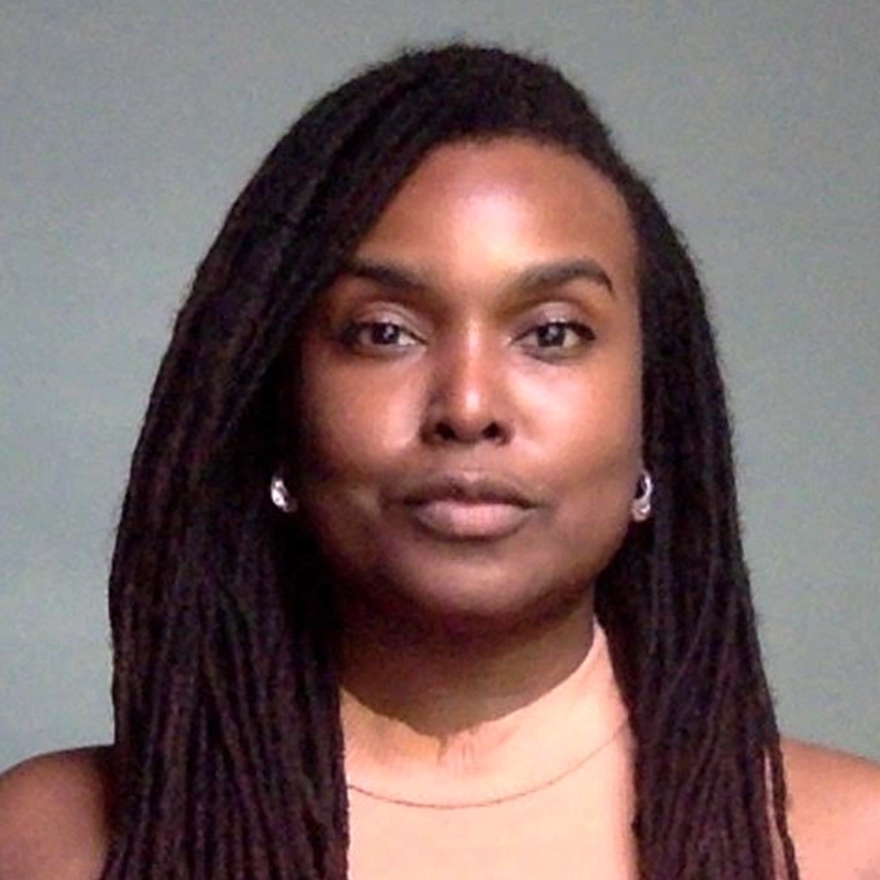 A person with long, dark, tightly coiled hair styled in locs, wearing a sleeveless beige top and small hoop earrings, looks directly at the camera with a neutral expression. The background is plain and grayish-green. This striking portrait captures Lori Parquet’s poised presence.