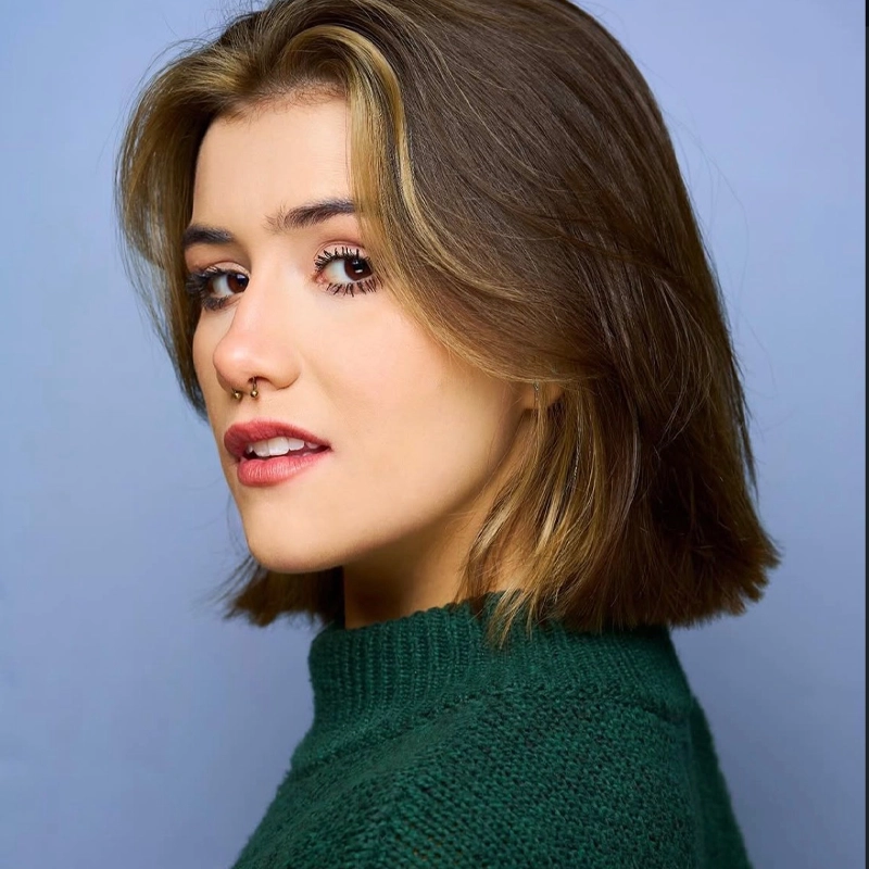 A young woman with straight, shoulder-length brown hair with blonde highlights looks over her shoulder. She wears a green sweater, has a septum piercing, and subtle makeup. The background is a solid light blue, perfectly complementing Audrey Loverro’s striking style.