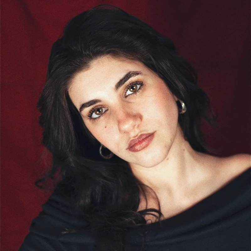 A woman with long dark hair and hazel eyes gazes at the camera with a neutral expression. She wears silver hoop earrings and a dark off-the-shoulder top. The background is deep red, and soft lighting highlights her face and shoulders.