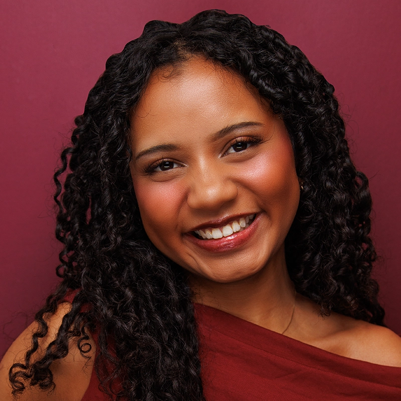 A smiling woman with long, curly dark hair looks directly at the camera. She wears a dark red top against a matching solid-colored background.