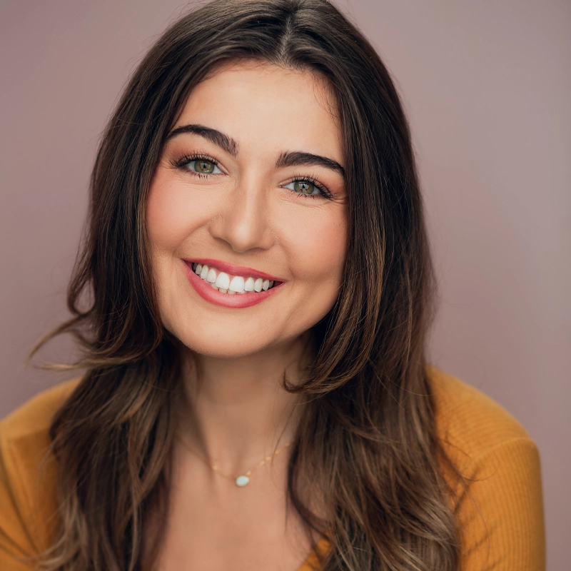 A woman with long brown hair, hazel eyes, and light skin smiles at the camera. She is wearing a mustard yellow top and a delicate necklace with a small pendant. The background is a soft, blurred mauve color.