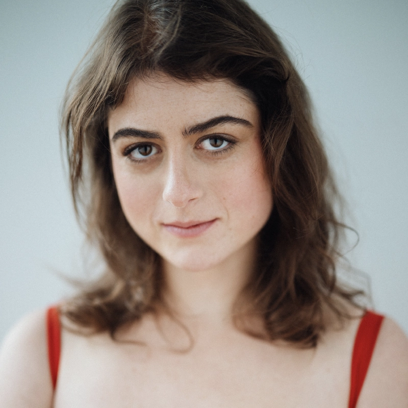 Ariella Silvina, a young person with wavy brown hair and fair skin, wears a red top, looking directly at the camera with a soft, subtle smile. The background is plain and light-colored, drawing focus to their facial expression.