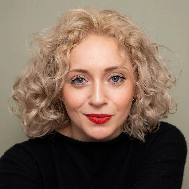 Celeste Zwingel, a woman with pale skin, curly blonde hair, and blue eyes, looks at the camera, smiling gently. She is wearing a black top and bright red lipstick. The background is plain and light-colored.