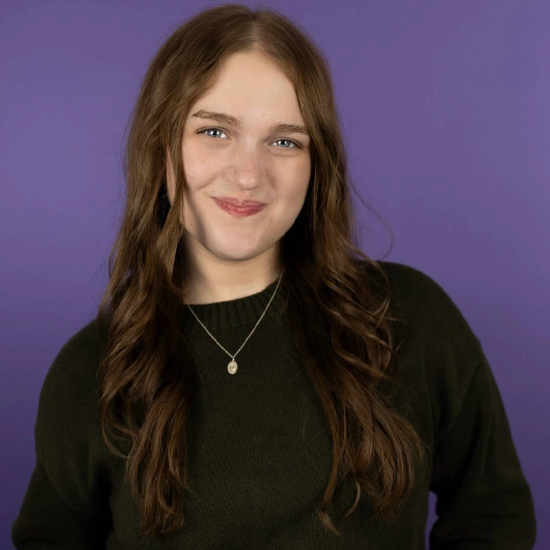 A young person with long, wavy brown hair wears a dark sweater and a pendant necklace, smiling softly at the camera. The background is a solid purple color.