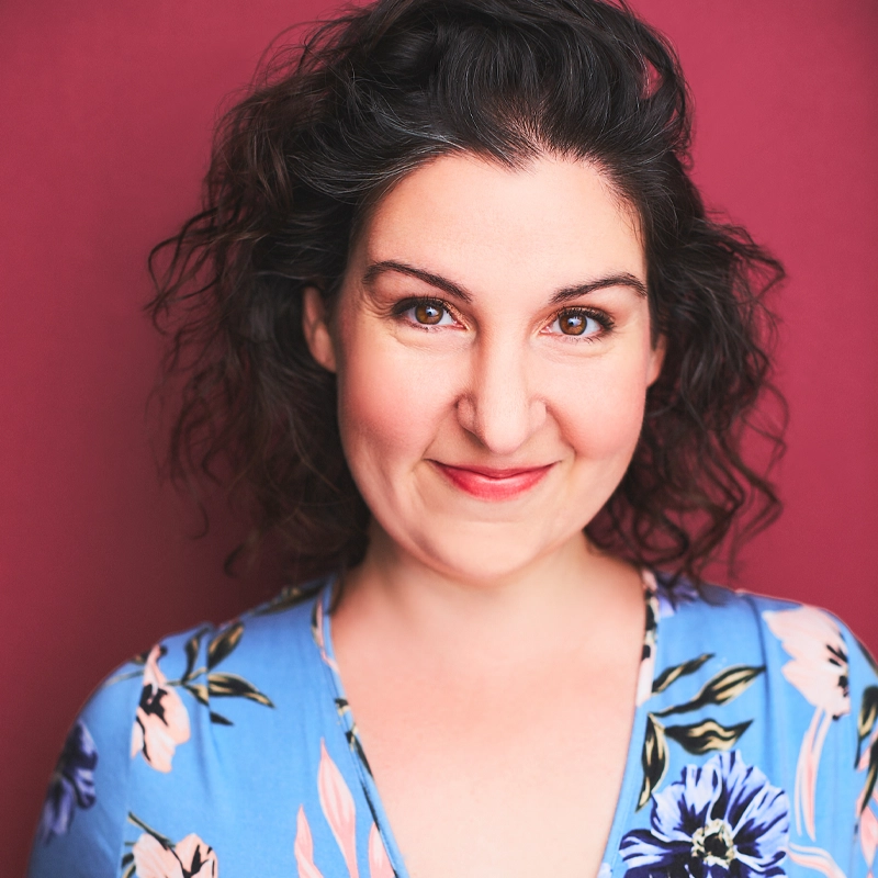 A woman with curly dark hair smiles softly at the camera. She wears a blue floral top and has natural makeup. The background is a solid, muted pink. Her expression is warm and approachable, perfectly capturing Eva Marie Mastrangelo’s inviting charm.