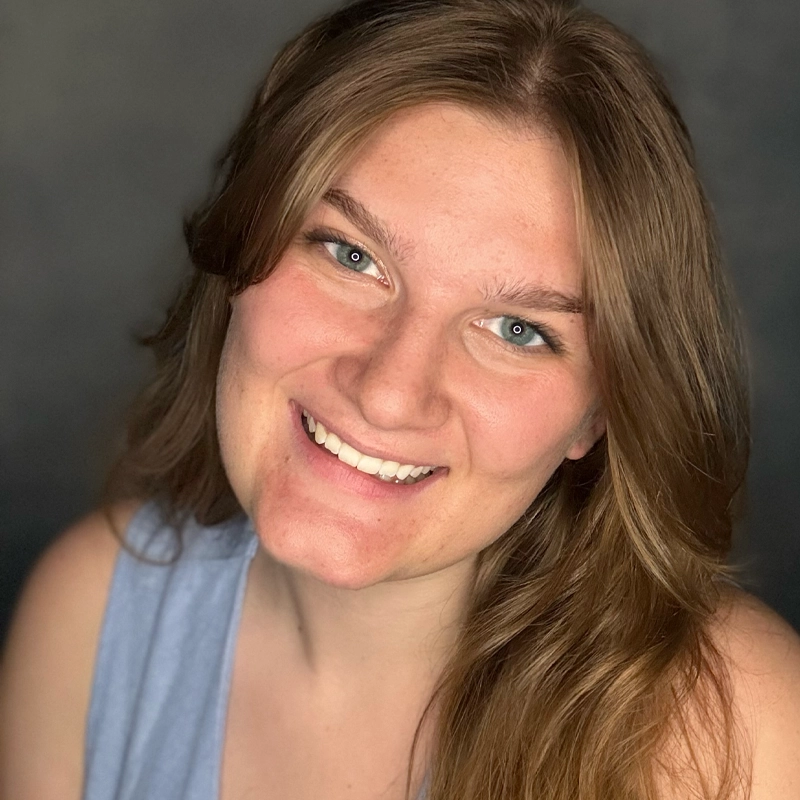 A young woman with long, light brown hair smiles at the camera. She wears a sleeveless, light blue top. The background is dark and softly blurred, making Brooklyn Mauch’s face the clear focal point of the image.