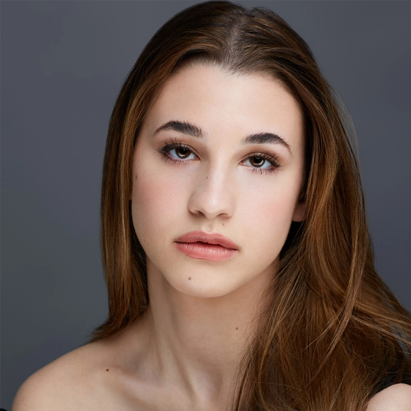 Young woman with long brown hair and fair skin looks directly at the camera with a neutral expression. Maya Levinson wears natural makeup, defined eyebrows, and no visible jewelry. The background is a plain, soft gray, creating a calm and focused portrait.