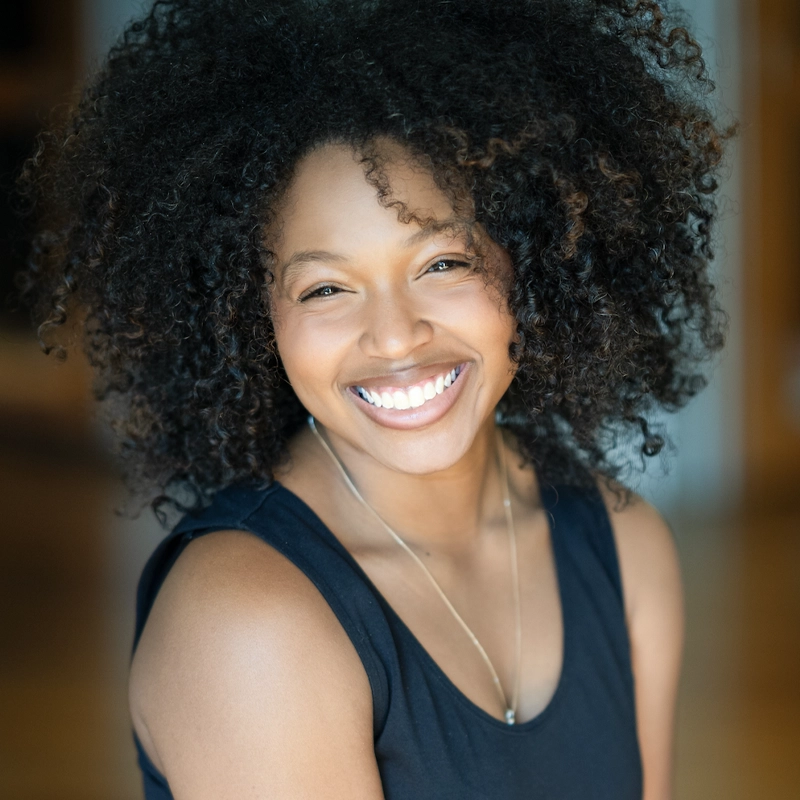 Azaria Imani McCants, a woman with voluminous curly hair and a big, bright smile, looks directly at the camera. She wears a sleeveless black top and a delicate necklace. The background is softly blurred, drawing focus to her cheerful expression.