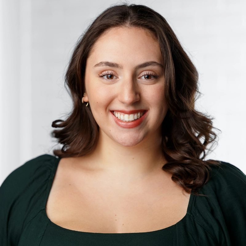 Julie McConnell, a young woman with wavy brown hair, smiles at the camera. She wears a dark green top with a square neckline and small gold hoop earrings. The background is softly blurred, featuring subtle white and light gray tones.