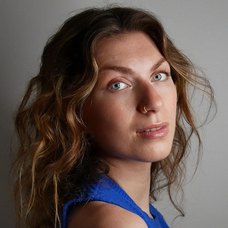 Celeste Zwingel, with wavy, light brown hair and blue eyes, looks over her shoulder at the camera. She wears a blue sleeveless top and a small nose ring. The background is plain and gray, highlighting her natural features and soft expression.
