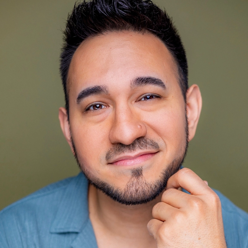 A person with short dark hair and a trimmed beard smiles gently, tilting their head slightly and touching their chin with one hand. Wearing a teal shirt, Delano Mendoza is posed against a plain olive green background.