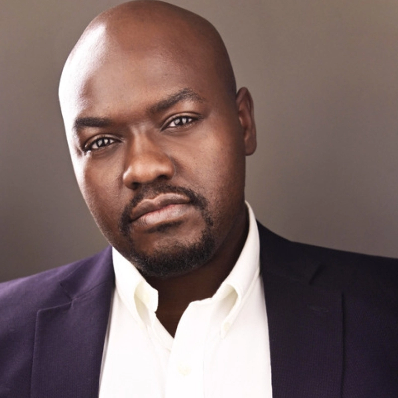 A man with a shaved head and short goatee, identified as Manny Milfort, wears a dark blazer over a white collared shirt and looks directly at the camera with a serious expression against a neutral background.
