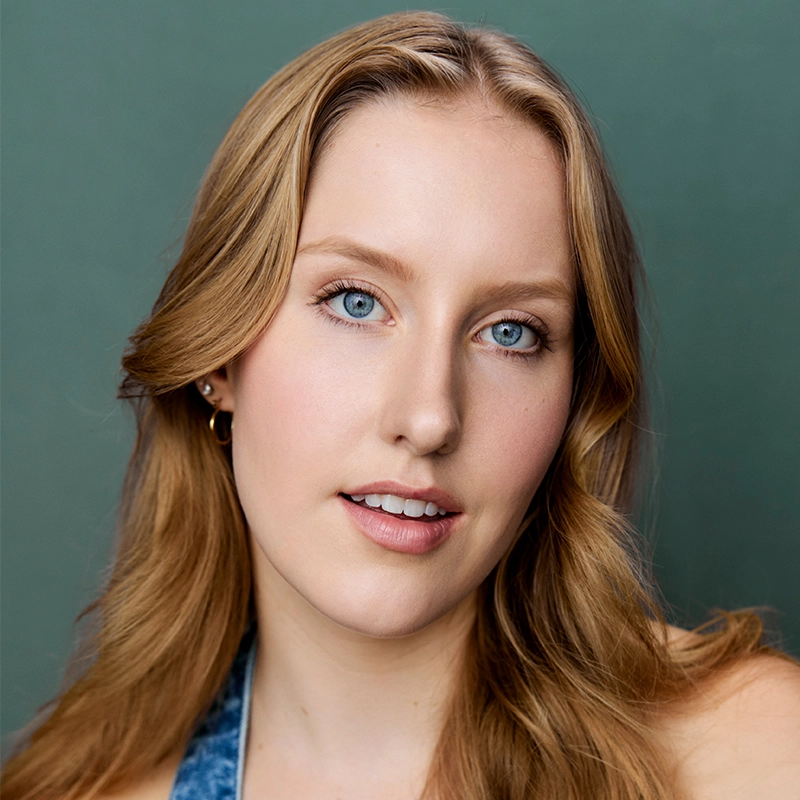 A young woman with long, wavy blonde hair and blue eyes looks at the camera. She wears natural makeup, small hoop earrings, and a blue top. The background is a soft green, and the lighting is even, highlighting Mimi Pietila’s clear skin and gentle expression.