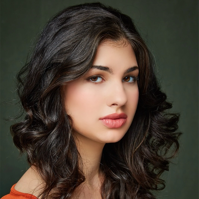 A young woman, Maya Levinson, with long, wavy dark brown hair gazes softly at the camera. She has fair skin, full lips, and brown eyes. She wears a rust-colored top, and the background is dark green, giving the image a classic portrait feel.