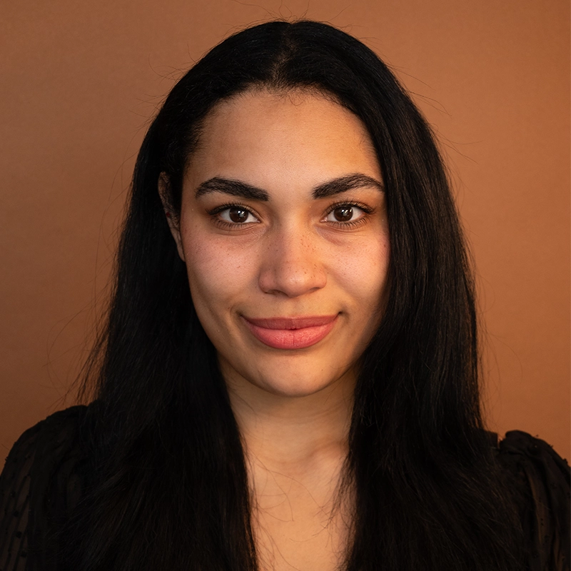 A woman with long straight black hair and medium brown skin looks directly at the camera, smiling slightly. She wears a black top and stands against a plain warm brown background. The soft, even lighting across her face highlights her features, reminiscent of portraits by Kendall Tubbs.