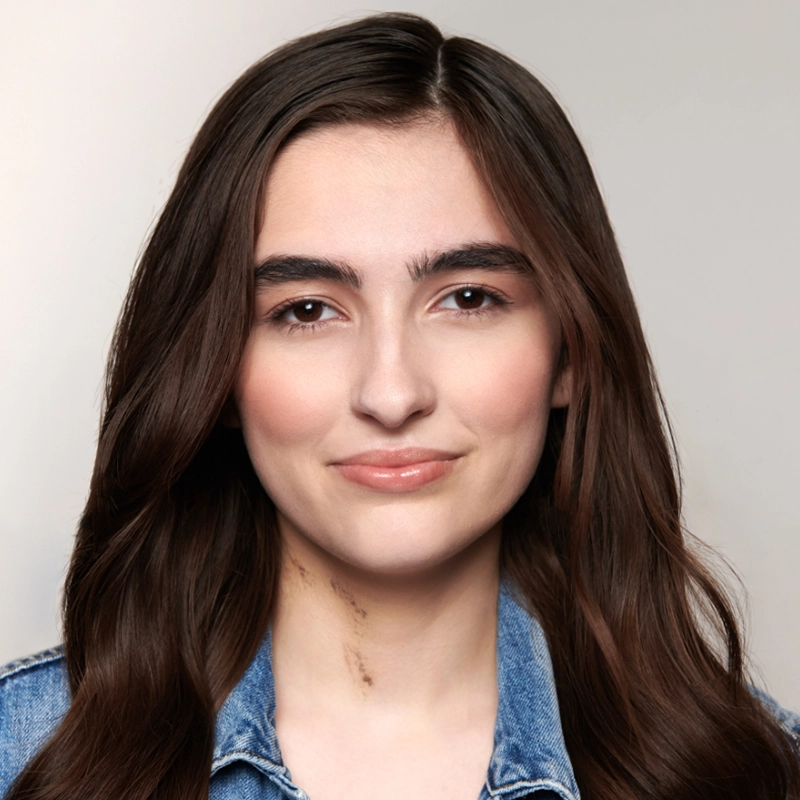 Young woman with long brown hair and fair skin wearing a blue denim jacket. She has natural makeup, thick eyebrows, and a slight smile. The background is plain and light-colored, keeping the focus on her face. A small scar is visible on her neck, giving Kelsey Valente’s features a distinctive touch.