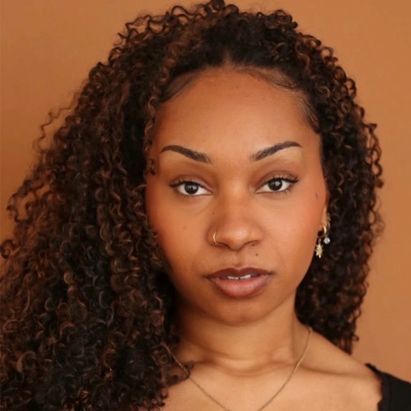 A woman with medium brown skin and curly dark brown hair looks directly at the camera. She has a small nose ring and wears earrings and a necklace. The background is a solid, warm brown color. Her expression is neutral and confident, embodying the poised presence of Kendall Tubbs.