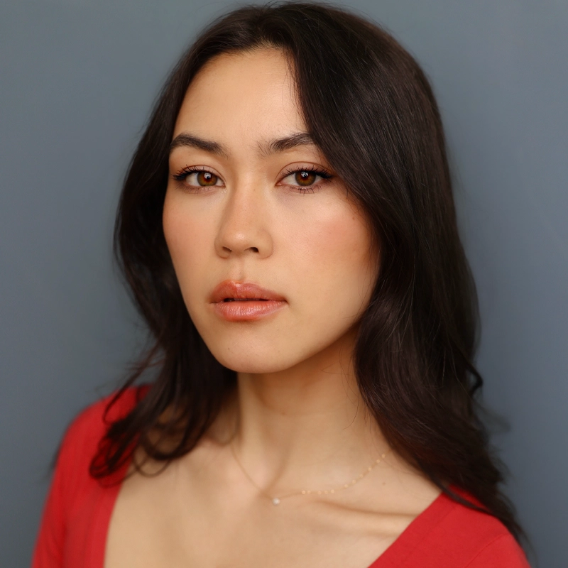 A woman with long, dark brown hair and fair skin gazes at the camera with a neutral expression. Malia Munley wears a red top and has subtle, natural makeup with peach lipstick. The background is plain gray, and soft lighting highlights her facial features.