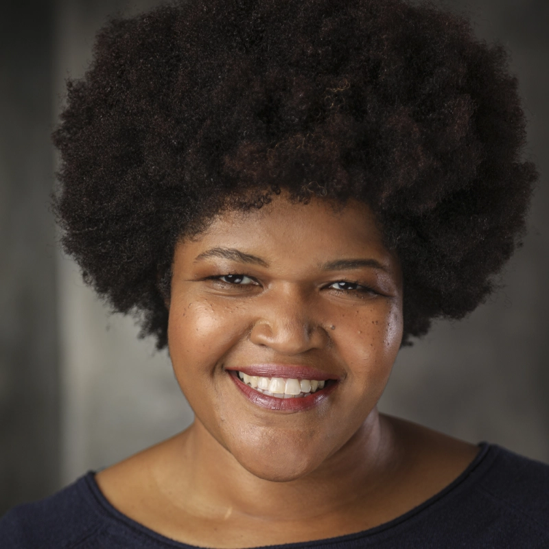 Smiling person with a dark curly afro hairstyle, wearing a dark shirt, faces the camera against a blurred gray background. With warm brown skin and expressive eyes, Matiwonesa Munyaradzi exudes warmth and friendliness through their natural look.