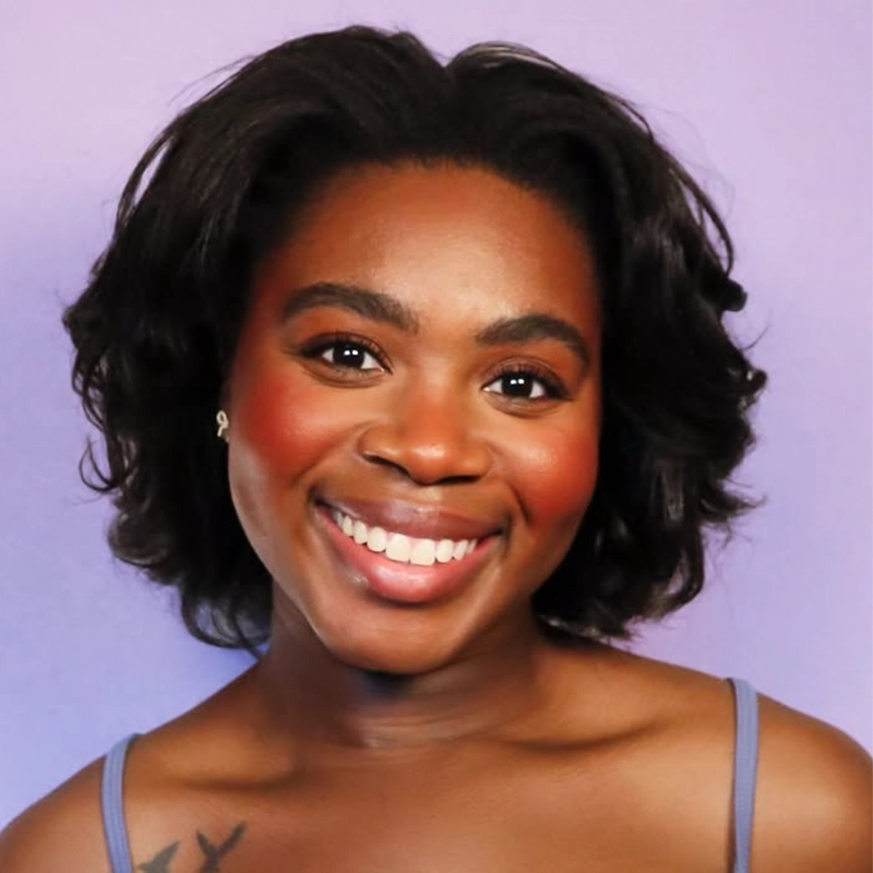 A woman with short, wavy black hair smiles warmly. She has glowing skin, peach blush, and natural makeup. She wears a strappy top and small earrings. The background is a solid pastel purple. A tattoo is partially visible on her left shoulder, giving a subtle hint of personal style reminiscent of Kendall Tubbs’ confident elegance.