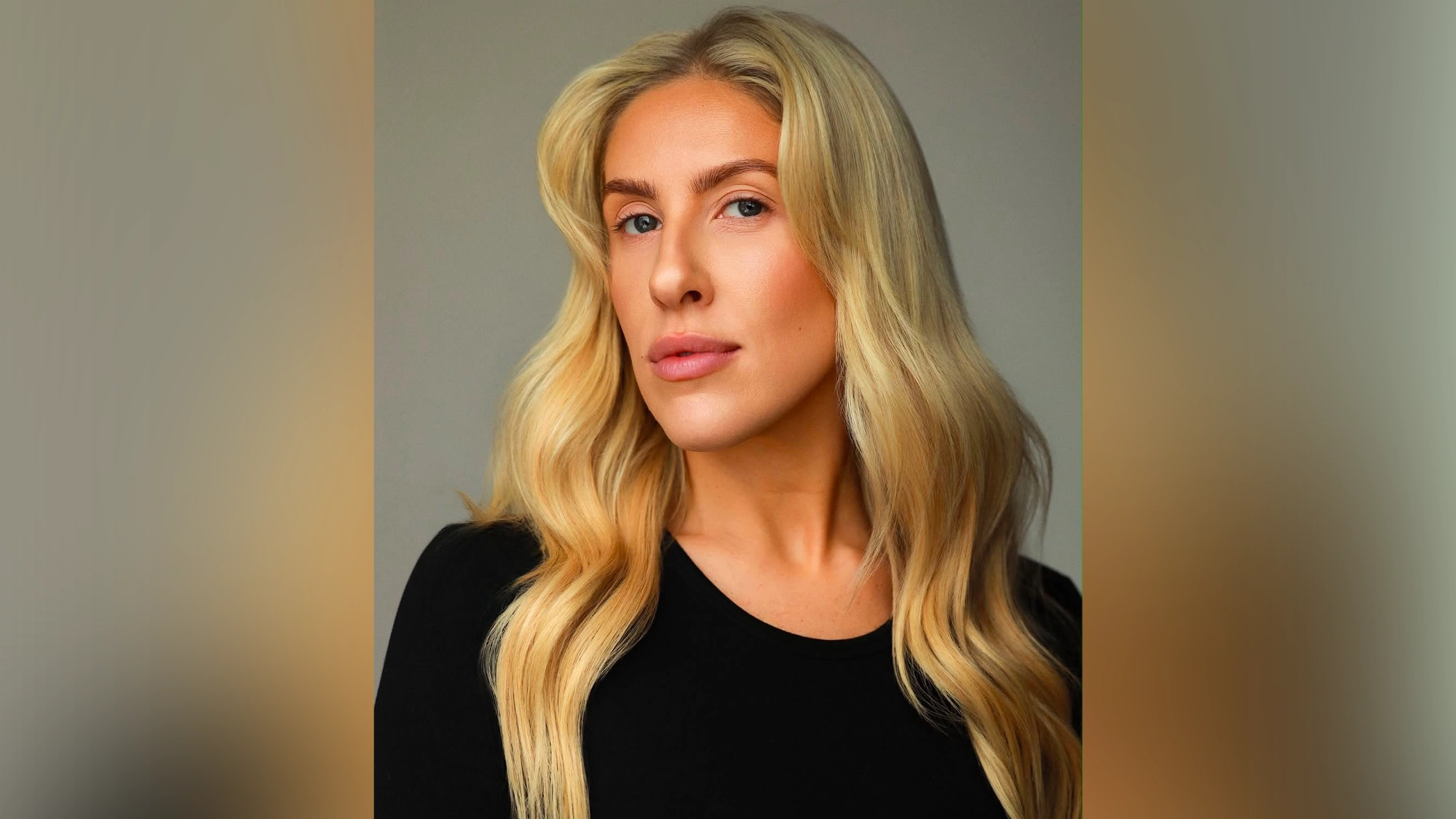 Natalie Paris - Site Image A person with long, wavy blonde hair and light skin gazes confidently at the camera. Reminiscent of Natalie Paris, they wear a simple black top. The background is a plain, neutral gray, emphasizing their facial features and minimal makeup.