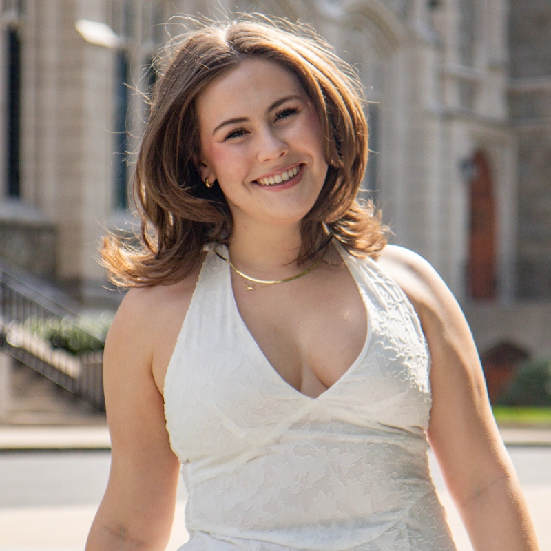 Eleanor Neary, a young woman with shoulder-length brown hair, smiles outdoors while wearing a sleeveless white dress with a deep V neckline. She stands in front of a stone building with arched windows, and the sunlight highlights her face and hair.