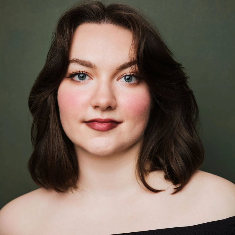 A young woman with fair skin, shoulder-length dark brown hair, blue eyes, and rosy cheeks is smiling softly. She wears natural makeup and an off-the-shoulder black top. The background is plain and dark olive green.
