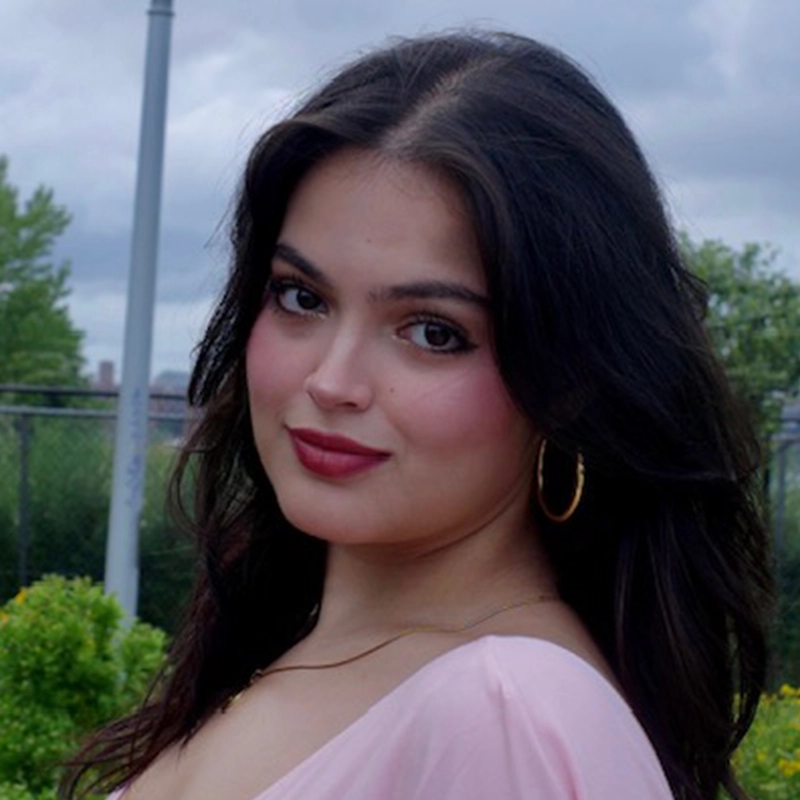 A young woman, Nicole Rodriguez, with long dark hair, gold hoop earrings, and a pink top smiles softly. She is outdoors, with a cloudy sky, greenery, and a fence in the background. Her makeup is subtle, with pink lipstick and defined eyebrows.
