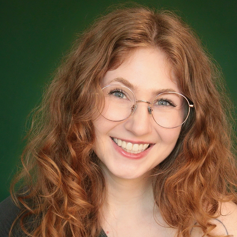 Smiling young woman, Alyssa Enita Stanford, with long, curly red hair and round glasses, wearing a dark top, in front of a dark green background. She is looking directly at the camera, showing her teeth.