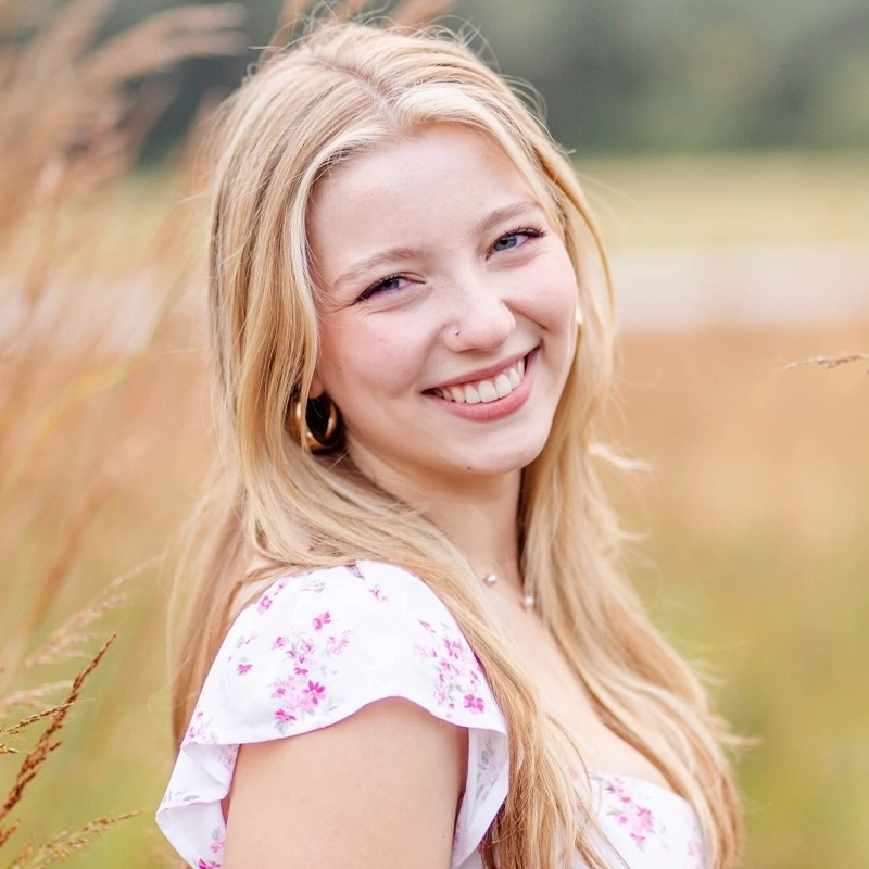 A young woman with long blonde hair smiles outdoors. Abigail Northrup is wearing a white dress with pink floral patterns and gold hoop earrings. The background is blurred with tall grass and greenery, and the lighting is natural and soft.
