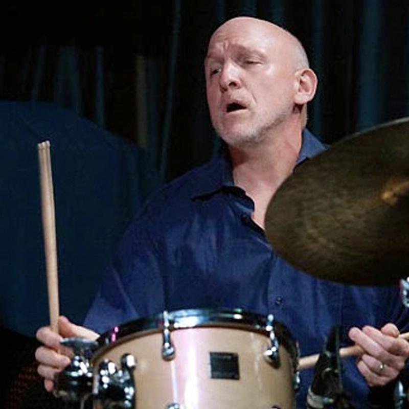A bald man with light skin, wearing a dark blue shirt, plays drums. He holds drumsticks in both hands and appears focused, with eyes half-closed and mouth slightly open. The drum set and cymbals are visible in the foreground, evoking the intensity of renowned drummer Adam Nussbaum. A dark backdrop is behind him.